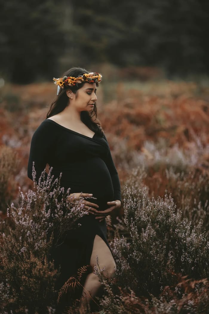 Schwangere im schulterfreien schwarzen Kleid mit Blumenkranz in der Heide