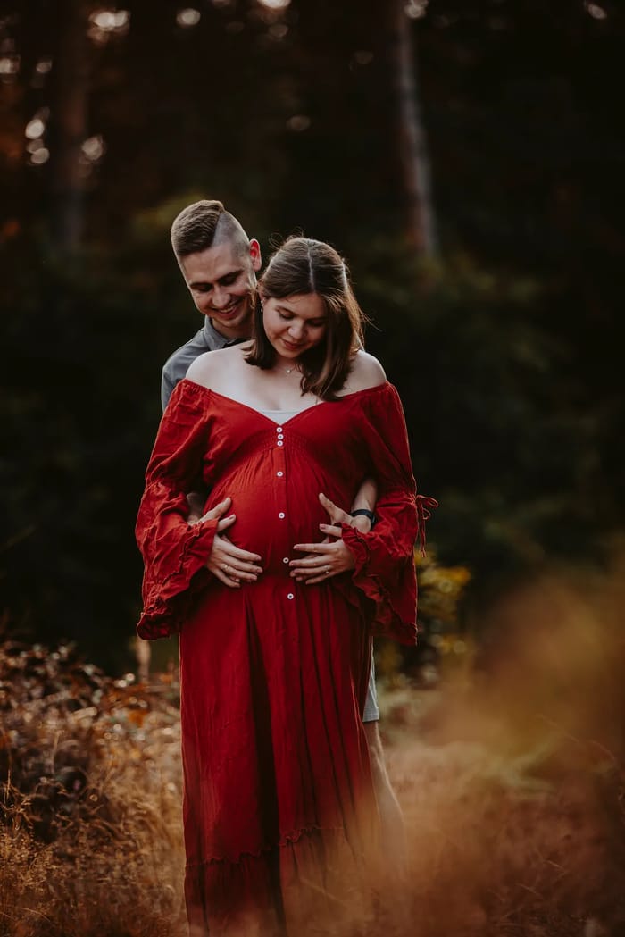 Werdende Eltern im Wald, sie im schulterfreien rostroten Kleid mit weiten Ärmeln