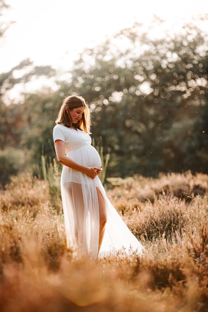 Schwangere im weißen Boho-Kleid im Gegenlicht auf einer Heidewiese